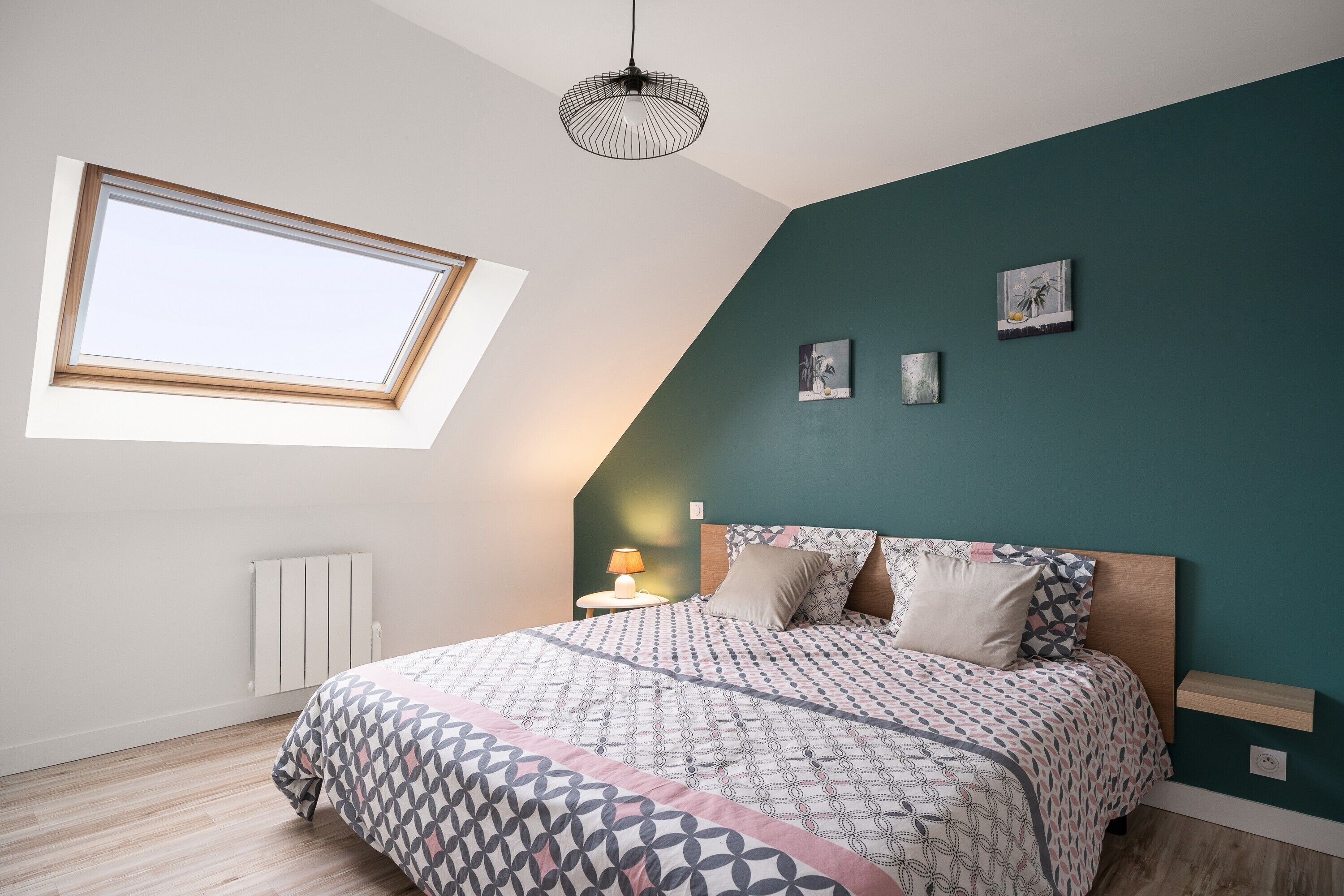 A bedroom with a sloped ceiling, a skylight, a bed with patterned bedding, a bedside lamp, and wall art.