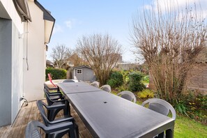 Une terrasse avec une longue table et des chaises, entourée d’un jardin où poussent différentes plantes, et un abri de jardin visible au fond.