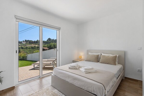 A bedroom with a bed, a nightstand, a lamp, a sliding glass door to a patio with chairs, and a view of greenery. - Holiday Home 'Refugio Das Vinhas' with Mountain View, Wi-Fi and Air Conditioning (Celorico de Basto)