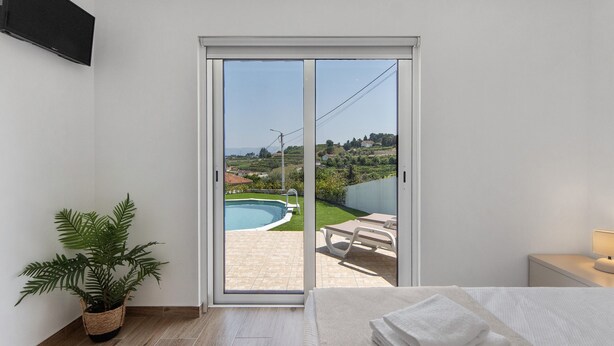 A bedroom with a bed, a bedside table, a lamp, a potted plant, and a sliding glass door leading to an outdoor area with a pool and lounge chairs. - Holiday Home 'Refugio Das Vinhas' with Mountain View, Wi-Fi and Air Conditioning (Celorico de Basto)