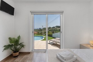 Ein Schlafzimmer mit Bett, Nachttisch, Lampe, Topfpflanze und einer Glasschiebetür, die zu einem Außenbereich mit Pool und Liegestühlen führt.