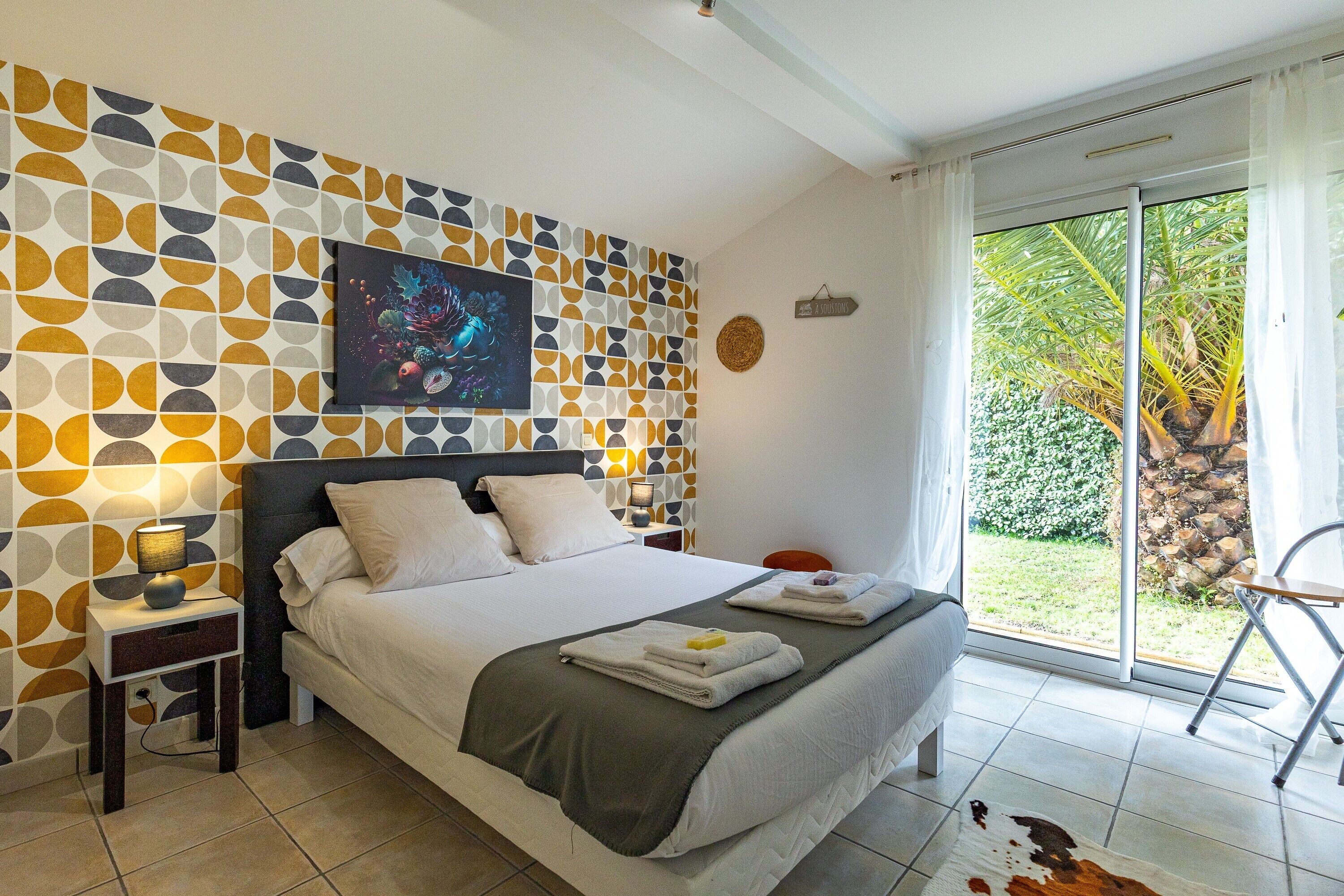 Une chambre à coucher comprenant un lit, une table de chevet, une œuvre d’art murale, une fenêtre donnant sur un espace verdoyant et une porte.