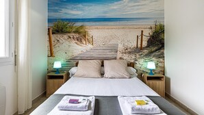 A bedroom with a beach view, two bedside tables with lamps, a bed with two pillows, and a wooden bench.