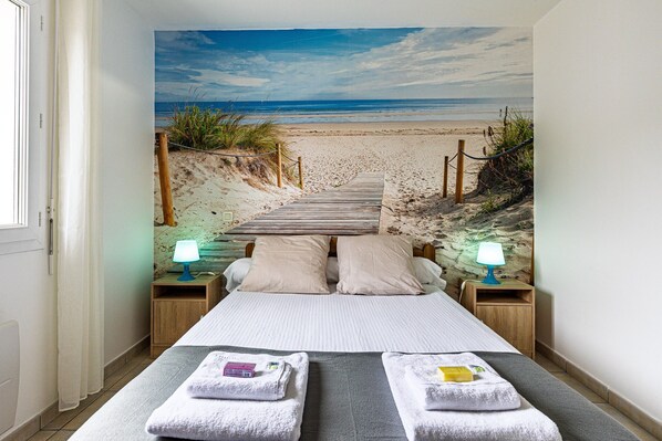A bedroom with a beach view, two bedside tables with lamps, a bed with two pillows, and a wooden bench.