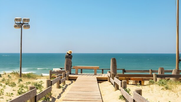 A wooden boardwalk leading to a bench overlooking a beach with clear skies.