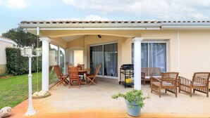 A covered patio with a table and chairs, a potted plant, and a grill cart.