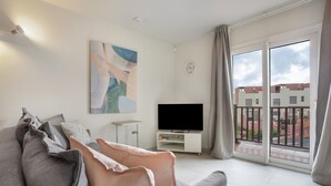 A modern living room with a sofa, a flat-screen TV, a painting, and a balcony with a view of buildings.