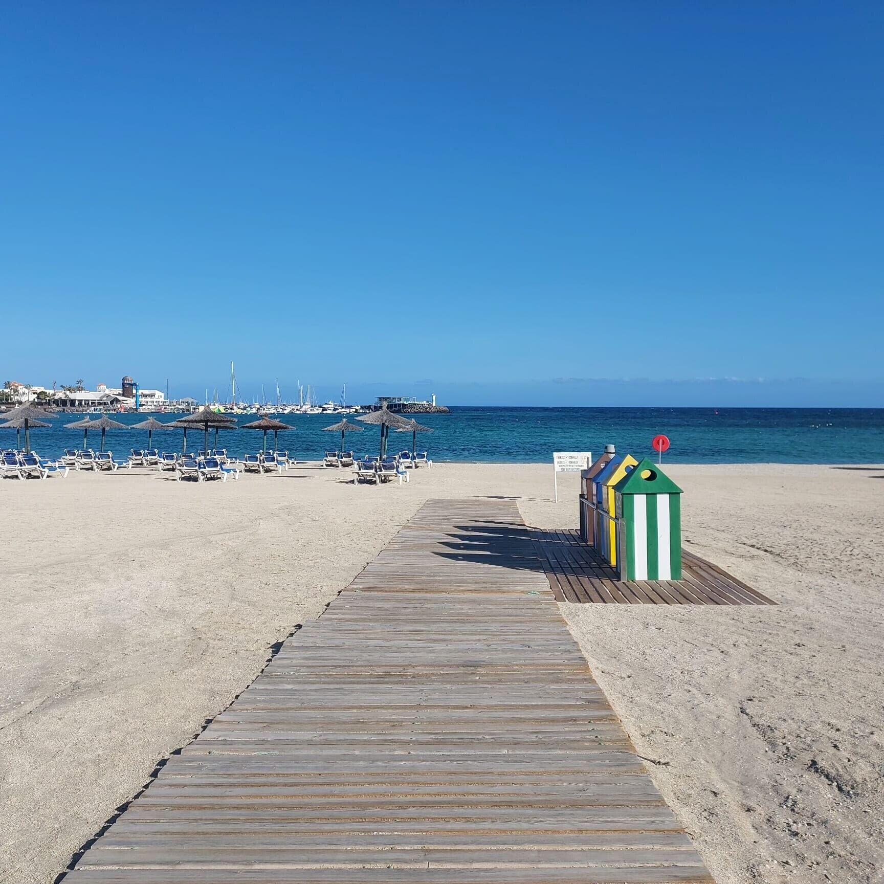 A wooden boardwalk leading to a beach with beach chairs and umbrellas, a small green and white structure, and a marina in the background.
