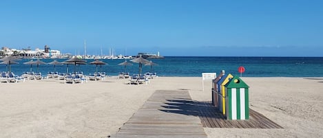 A wooden boardwalk leading to a beach with beach chairs and umbrellas, a small green and white structure, and a marina in the background.