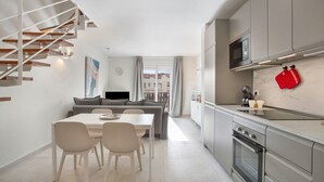 A modern kitchen with a built-in oven, a dining area with a table and chairs, a grey sofa, and a staircase with wooden steps.