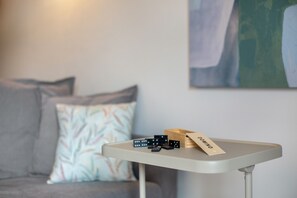 Une petite table avec des jeux de dominos et une carte portant l’inscription « DOMINO » dans une pièce où se trouvent un canapé gris et un mur orné d’œuvres d’art abstraites.