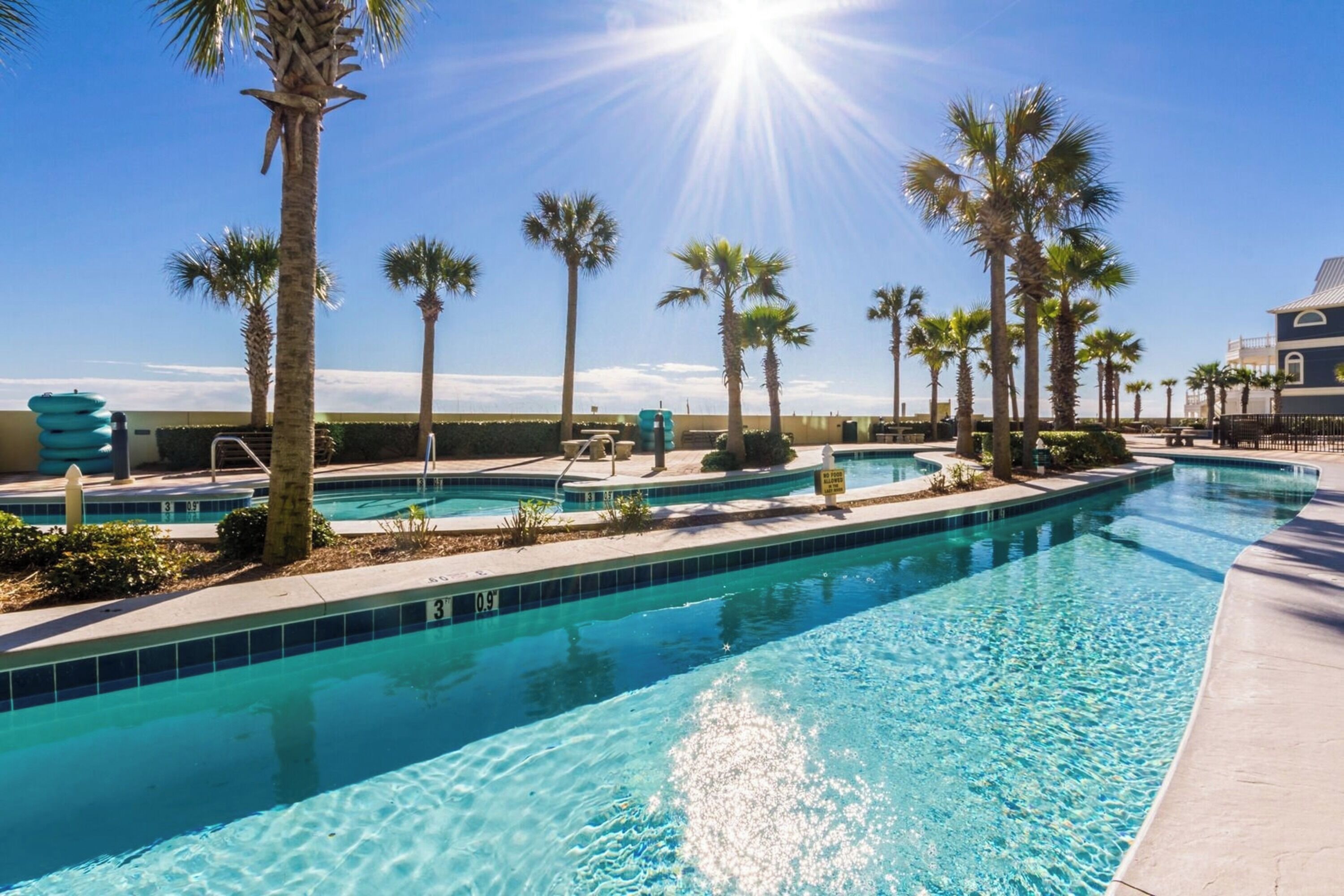 A swimming pool with clear blue water, surrounded by palm trees and a building in the background.