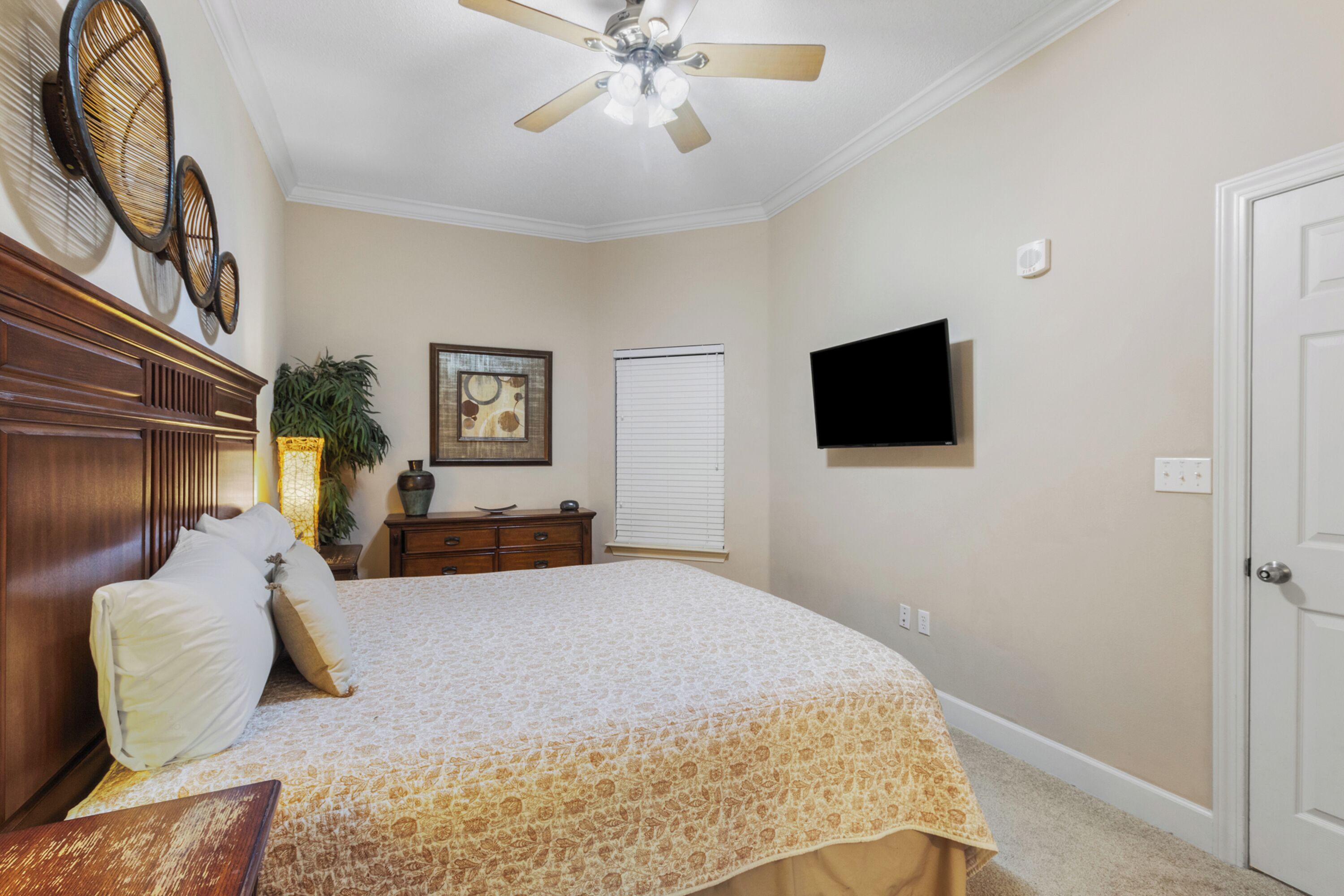 A bedroom with a bed, a ceiling fan, a television, a door, and a window with blinds.