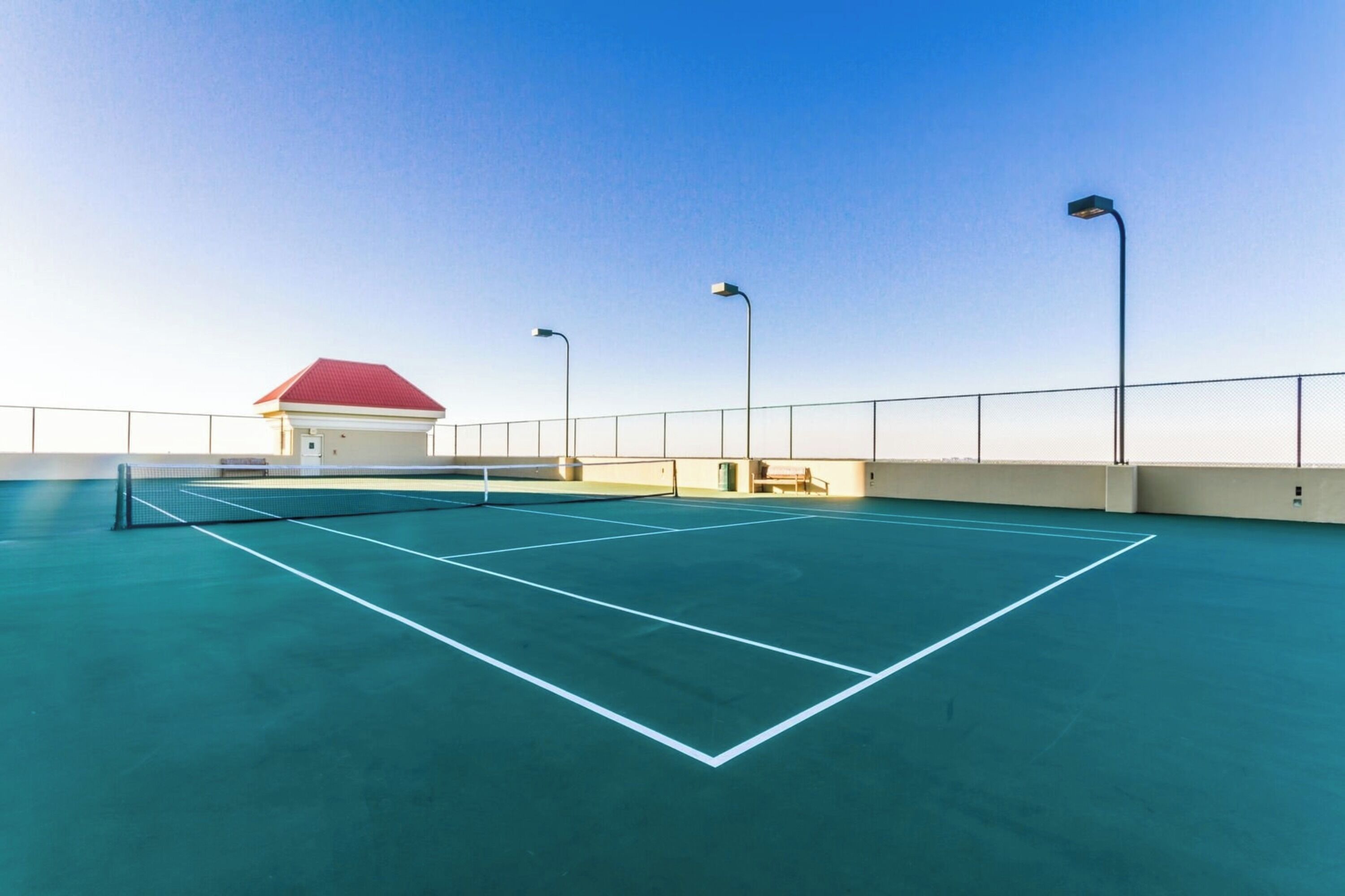 An outdoor tennis court with a net, surrounded by a low fence and three street lamps.