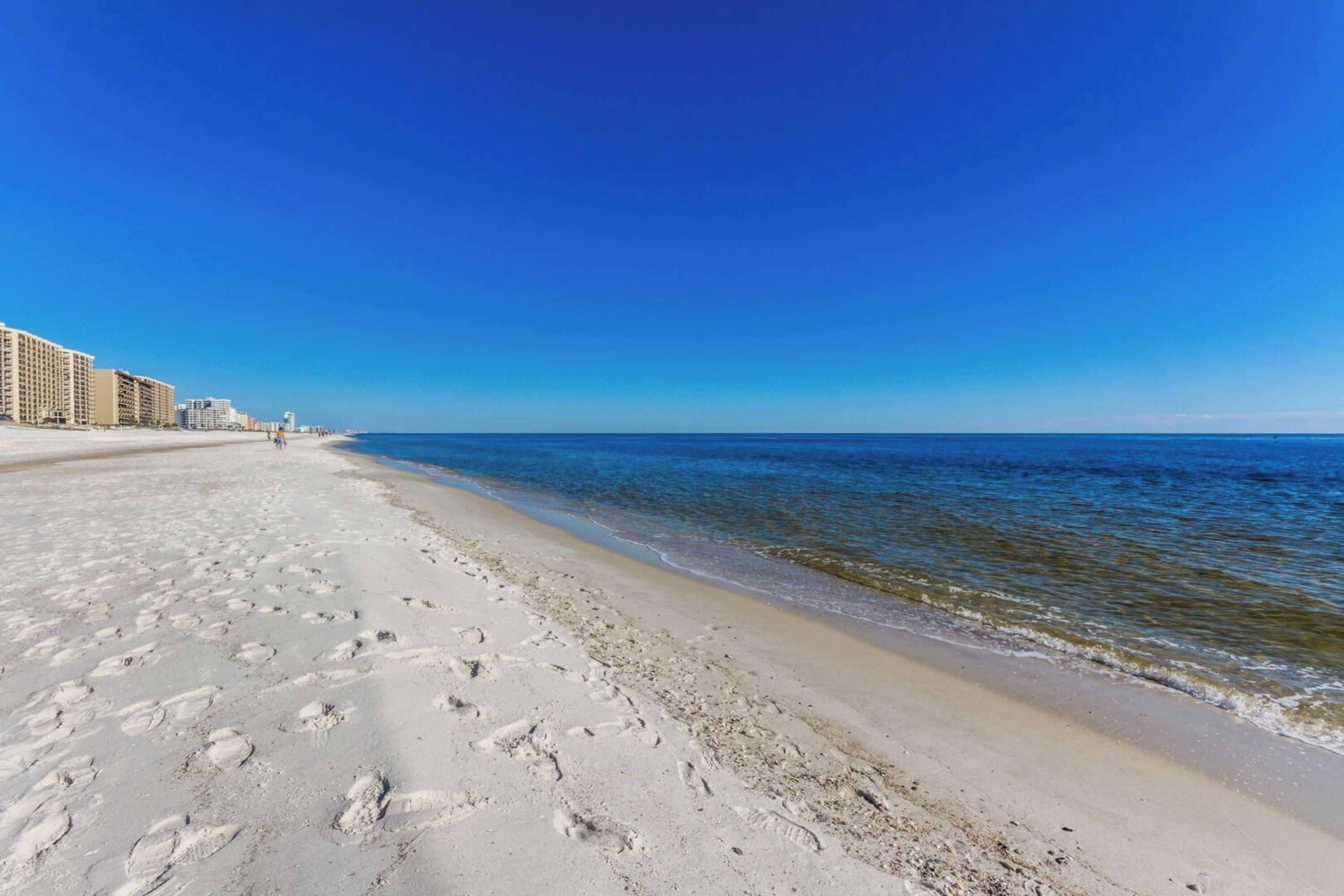 A beach with footprints in the sand, a clear blue sky, and buildings in the background.