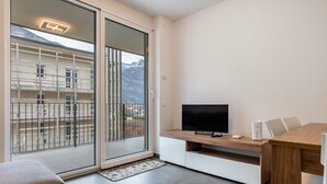 Een moderne kamer met een televisie, een houten eettafel en een glazen schuifdeur naar een balkon met uitzicht op de bergen.