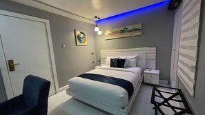A hotel room with a bed, bedside table, chair, and artwork on the wall. - The Stallion (Abuja)