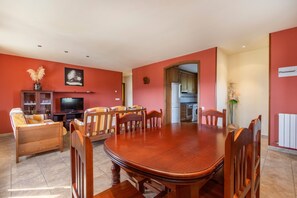 Zona pranzo con tavolo e sedie in legno, un divano, una televisione e una cucina sullo sfondo.