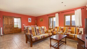 Sala de estar con sofá, una mesa de centro y televisor. Hay ventanas con cortinas, una puerta y un radiador.