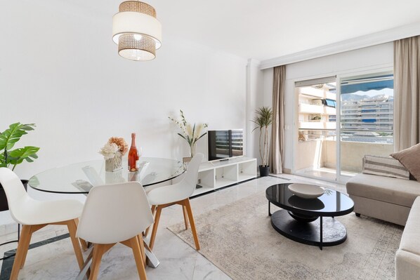 Ein modernes Wohnzimmer mit einem Glastisch, weißen Stühlen, einem schwarzen Couchtisch, einer Couch und einem Fernseher auf einem Lowboard.