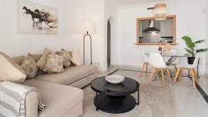A modern living room with a sofa, a round coffee table, a dining area with chairs, a kitchen with a mirror, and a potted plant.