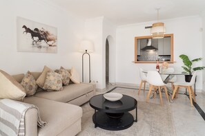 Ein modernes Wohnzimmer mit einer Couch, einem runden Couchtisch, einer Essecke mit Stühlen, einer Küche mit einem Spiegel und einer Topfpflanze.