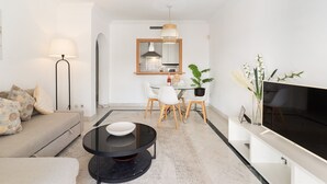 A modern living room with a sofa, a round glass coffee table, a TV, a dining area with a table and chairs, and a large mirror.