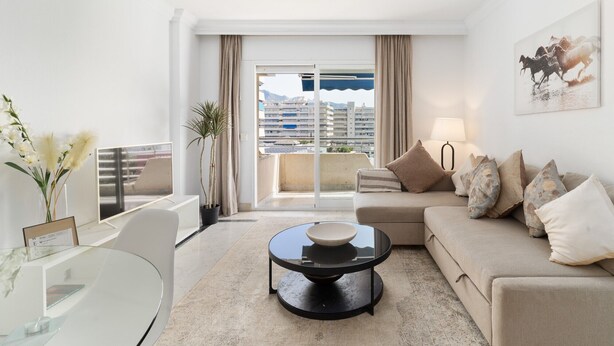 A modern living room with a sofa, coffee table, and a view of buildings through large windows.
