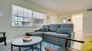 Ein Wohnzimmer mit grauem Sofa, einem runden Tisch mit einer Schale, einem Fenster mit Jalousien und einer Tür zu einem anderen Raum.