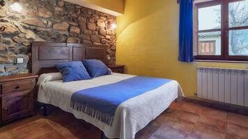 Un dormitorio con pared de piedra, cabecera de madera, cama con ropa de cama azul, mesita de noche y ventana con cortinas azules.