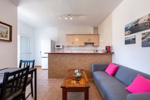Une cuisine moderne avec des meubles en bois, une table à manger avec des chaises, un canapé gris avec des coussins roses et une petite table sur laquelle se trouvent un bol et un verre.