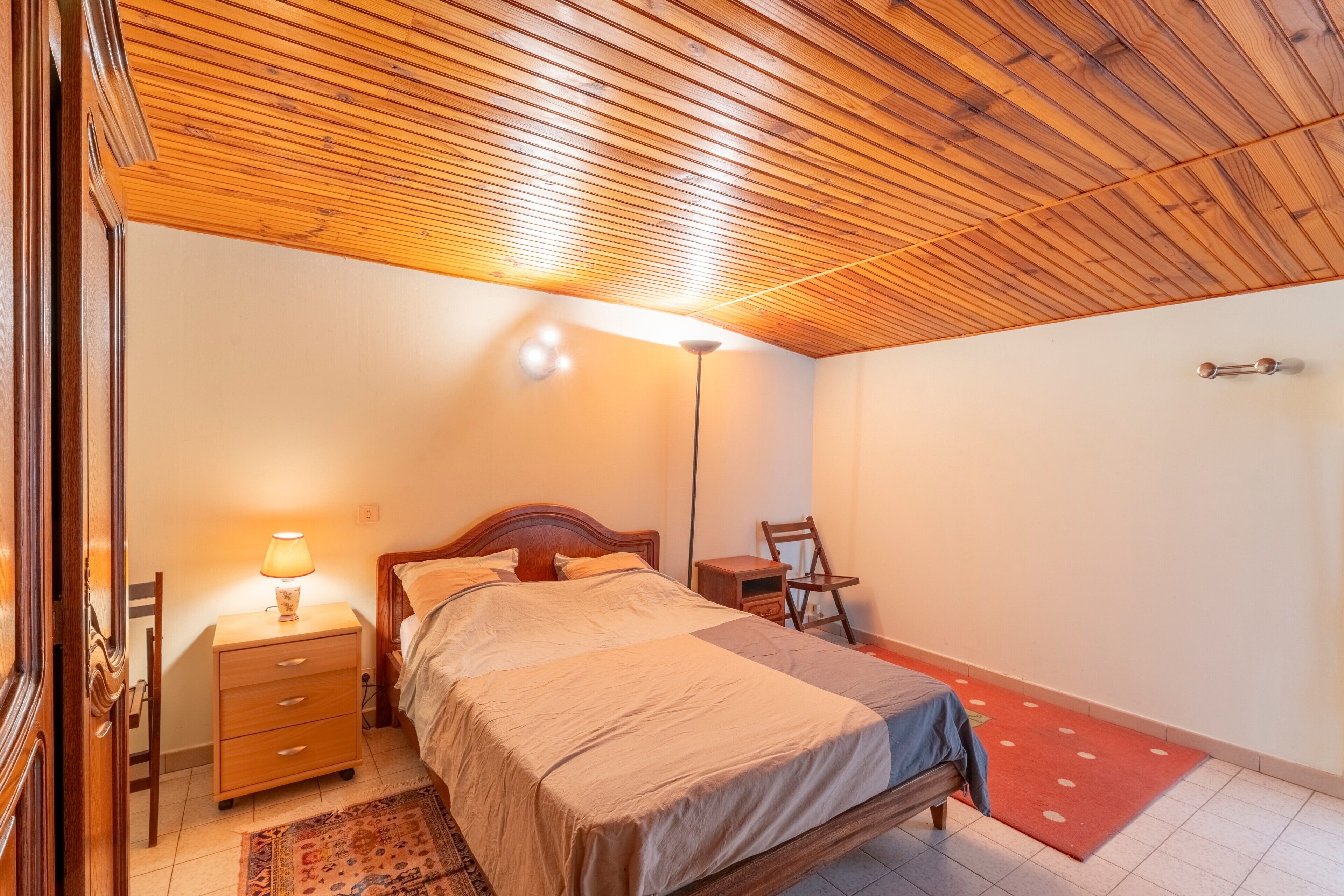 Una camera da letto con soffitto in legno, un letto, un comodino, una lampada, una sedia e un tappeto rosso.