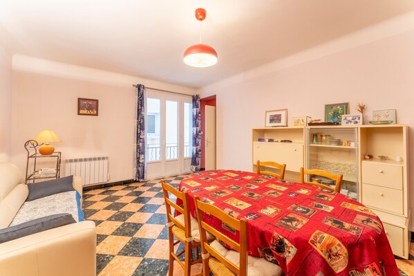 A room with a checkered floor, a dining table with chairs, a sofa, a radiator, a window with curtains, and a shelf with items.