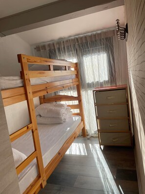 Ein Etagenbett mit Holzrahmen und weißer Bettwäsche, eine Kommode mit Schubladen und ein Fenster mit leichtem Vorhang.