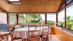 A sunroom with a wooden ceiling, large windows, a dining table with chairs, and a sofa.