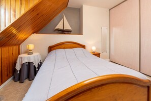 Um quarto com teto inclinado, cama com cabeceira de madeira, uma mesinha com abajur e decoração náutica de um veleiro na parede.