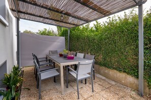 Um pátio externo com mesa e cadeiras, cercado por um viveiro e uma pérgula de metal.