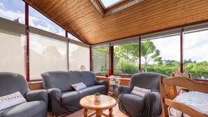A room with a wooden ceiling, large windows, a grey sofa, a wooden chair, and a small round table.