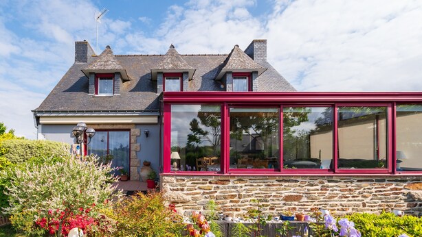 A stone house with a red-framed extension, a chimney, and a garden with potted plants.