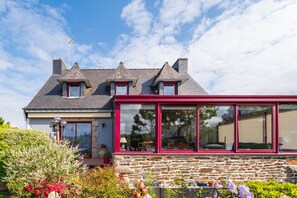 Uma casa de pedra com um anexo de estrutura vermelha, uma chaminé e um jardim com plantas em vasos.