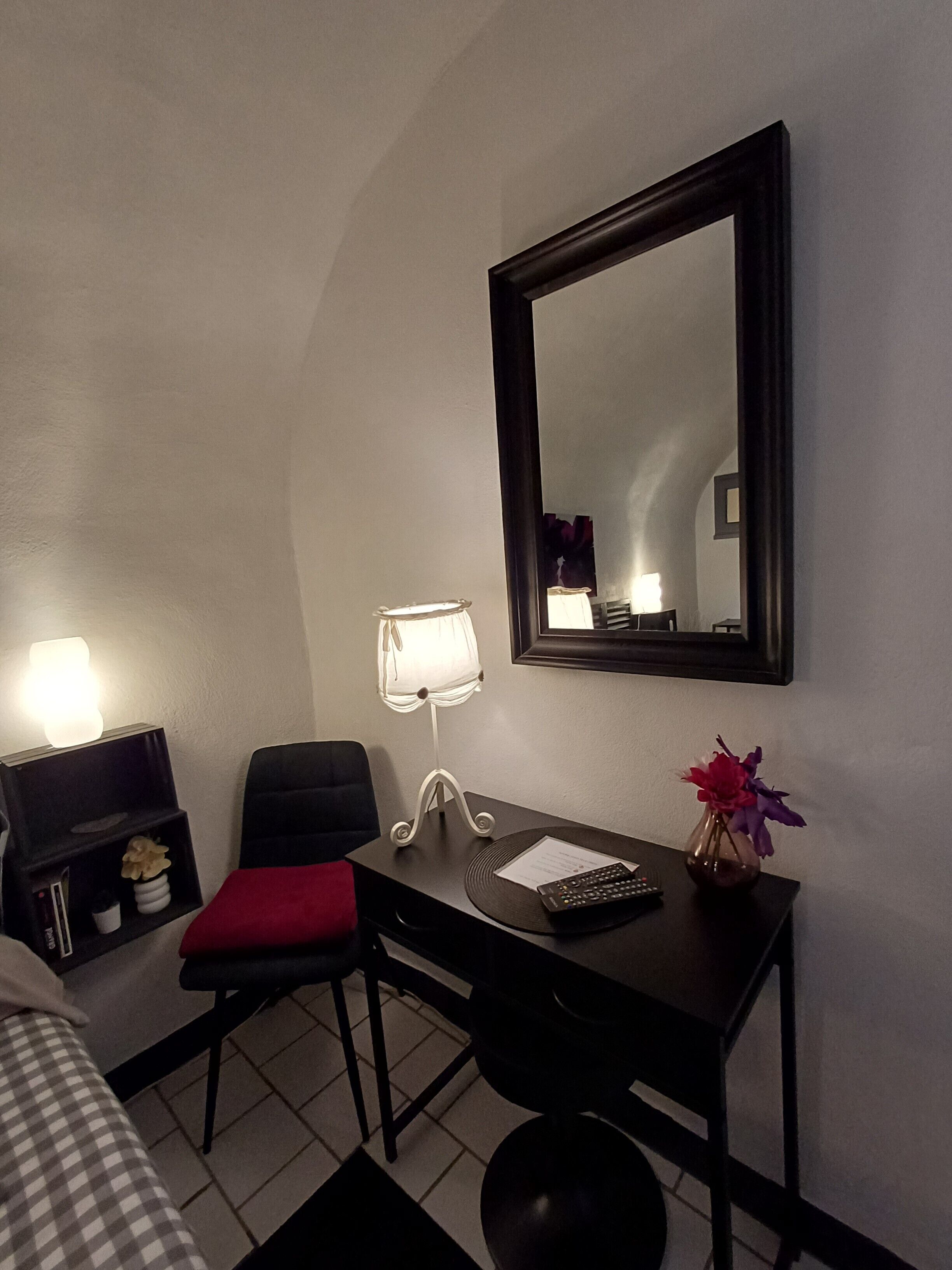 Una habitación de hotel con una cama, un escritorio con una silla, un estante pequeño, una lámpara, un espejo y un jarrón con flores.