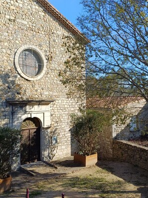 A stone building with a circular window, an arched entrance, and a small garden area with a planter and shrubbery.