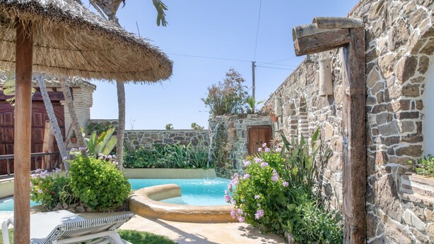 Un bâtiment en pierre avec une piscine, un parasol en chaume et un fauteuil de jardin blanc.