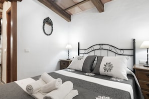 Ein ordentlich bezogenes Bett mit einem blumigen Überwurf, ein Nachttisch mit einer Lampe und ein an der Wand befestigter Spiegel.
