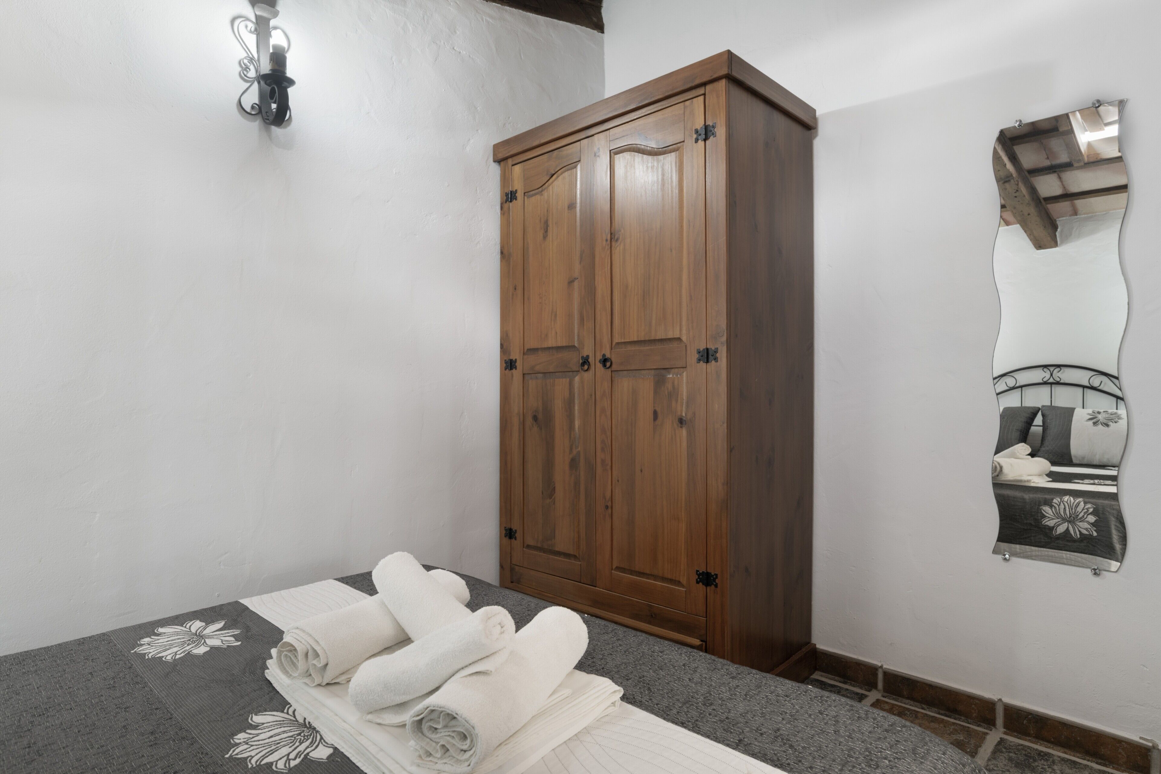 A bedroom with a wooden wardrobe, a bed with a floral-patterned bedspread, and a wall-mounted mirror.