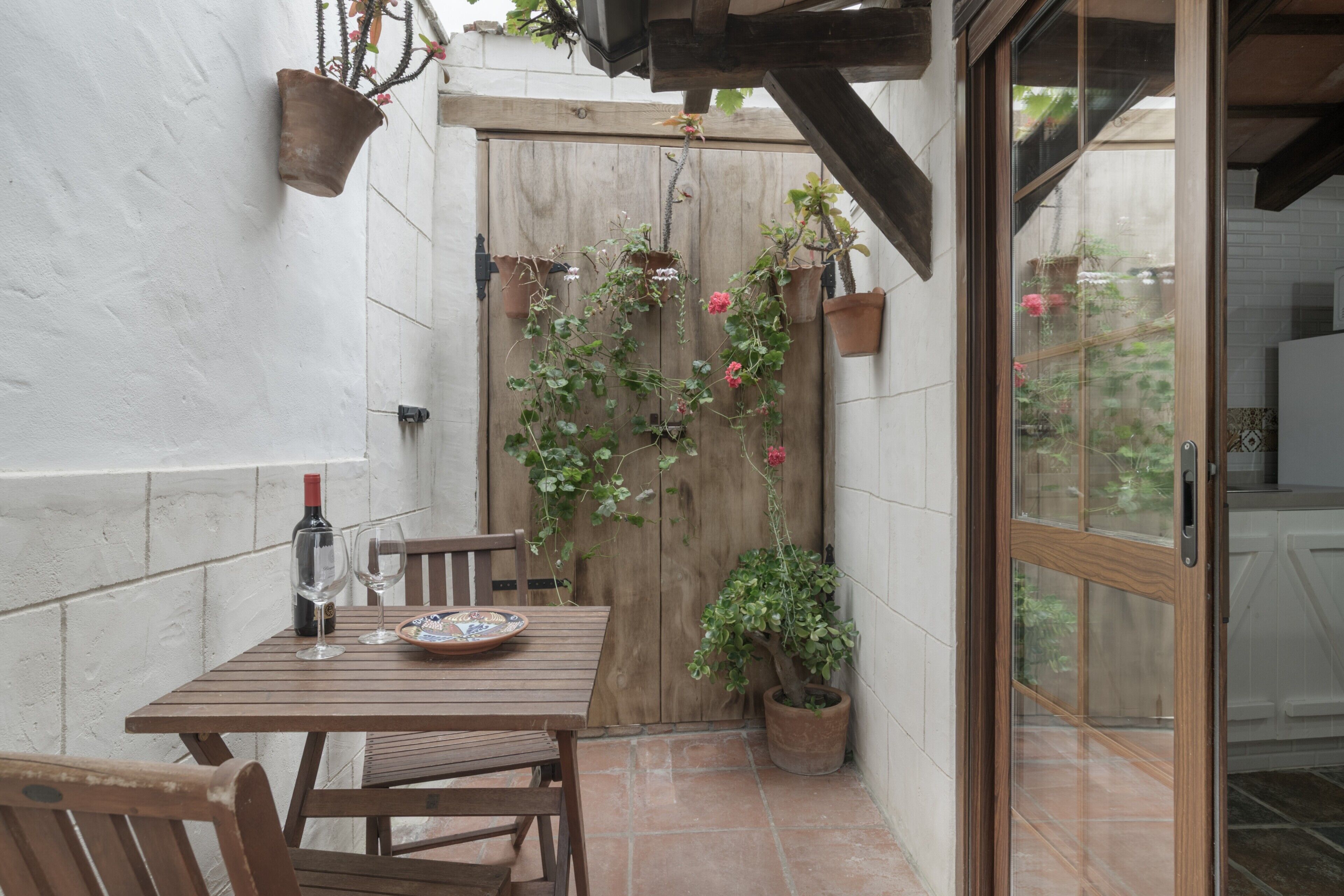 Ein Holztisch mit Stühlen, eine Weinflasche und Gläsern auf einer Terrasse mit Topfpflanzen und einer Holztür.