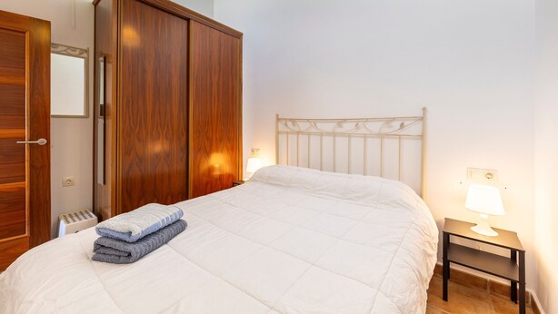 A bedroom with a bed, a wooden wardrobe, a bedside table with a lamp, and a wall-mounted light fixture.
