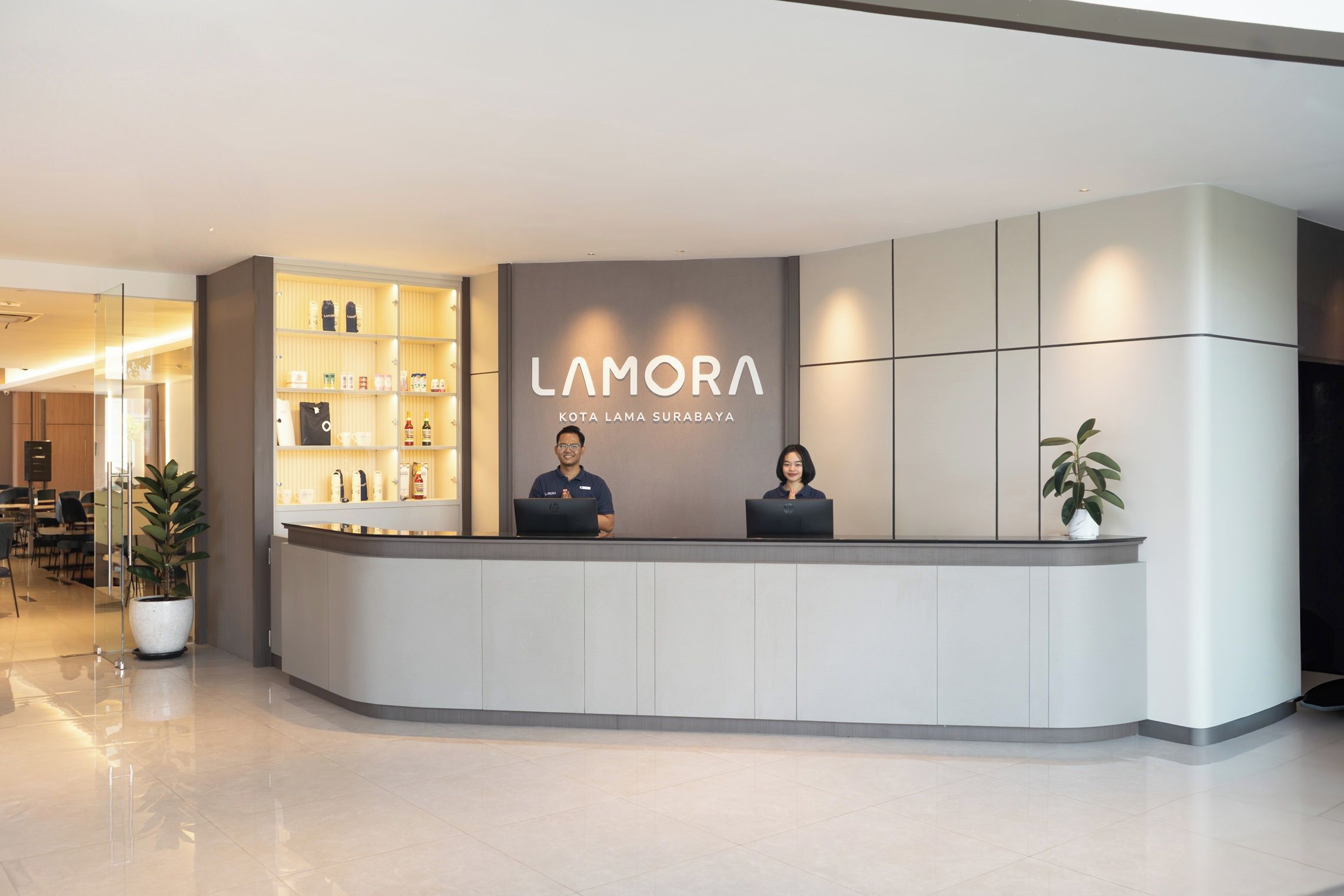A reception desk with a modern design, featuring a nameplate for 'LAMORA KOTA LAMA SURABAYA'. There are two staff members seated behind the desk, and a glass display case with various items on the left.