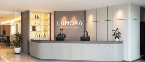 En reception med et moderne design, med en skilt med 'LAMORA KOTA LAMA SURABAYA'. To ansatte sidder bag disken, og der er en glasmontre med diverse genstande til venstre.