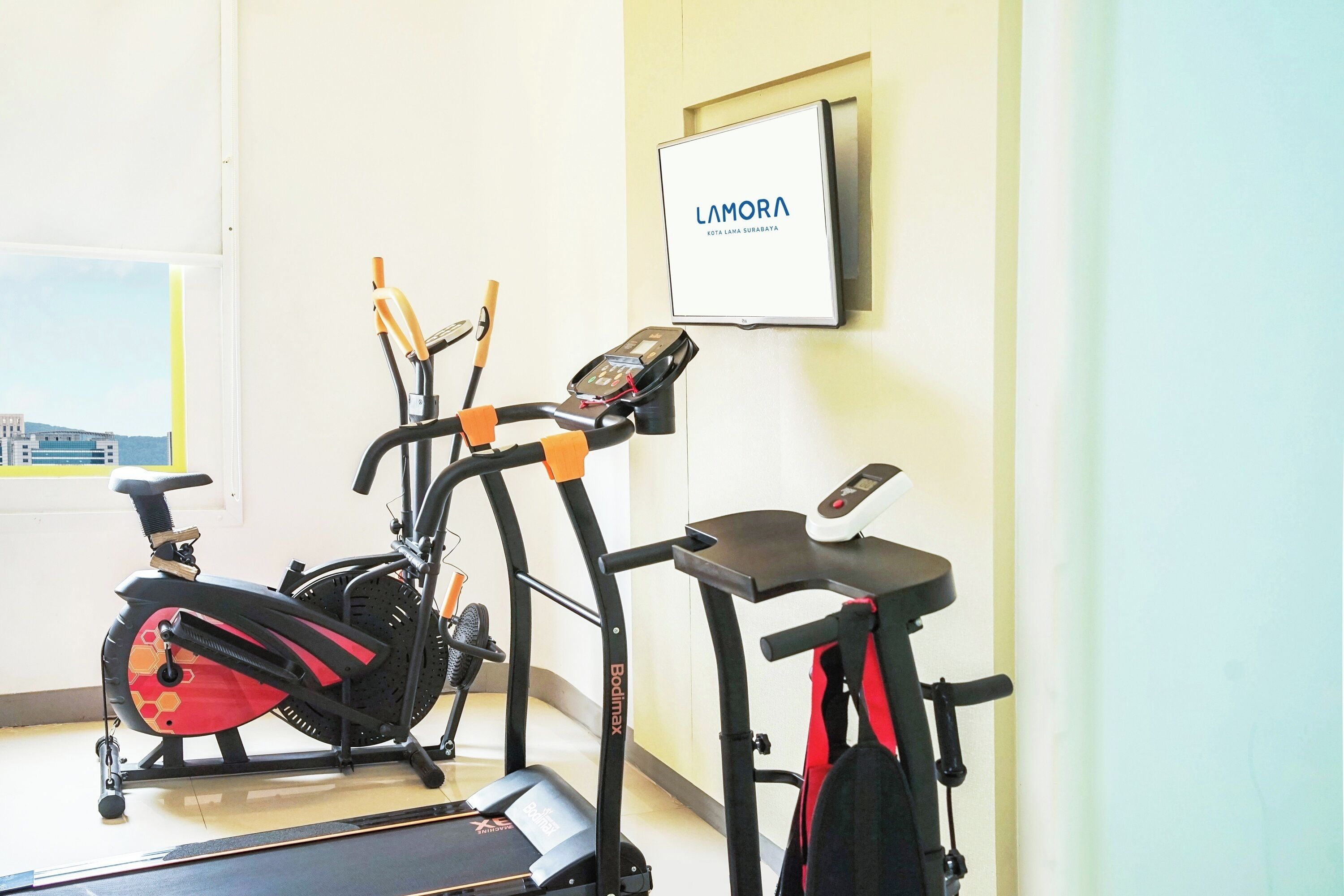 A stationary bike and treadmill in a room with a window and a wall-mounted TV displaying "LAMORA".